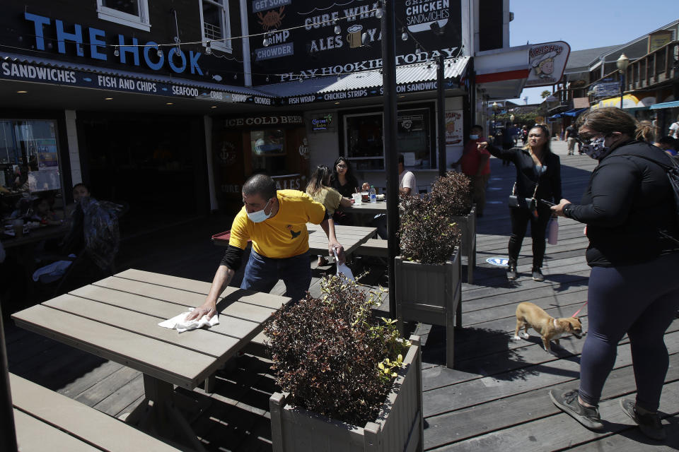 FILE - In this June 18, 2020, file photo a man wears a face mask while cleaning an outdoor dining table at The Hook at Pier 39, where some stores, restaurants and attractions have reopened, during the coronavirus outbreak in San Francisco. A new poll from The Associated Press-NORC Center for Public Affairs Research shows Americans becoming less concerned about infection and less supportive of restrictions. But with experts warning that hospitals in some states in the South and West will soon be inundated, the poll finds many Americans have not embraced reopening even as state and local officials have. (AP Photo/Jeff Chiu, File)