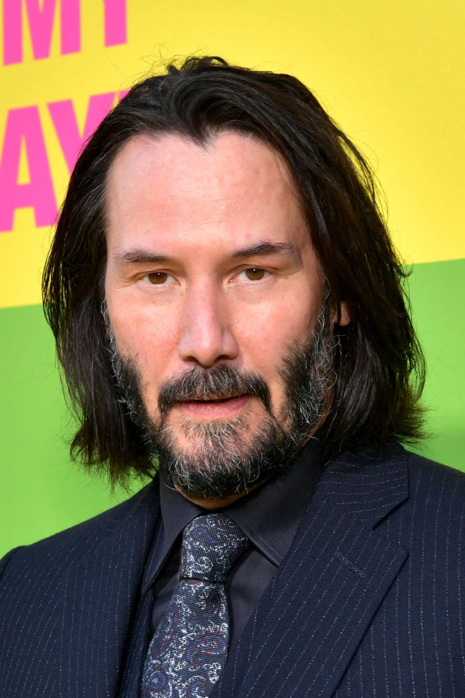 Keanu in a pin-striped suit and tie poses at event, smiling slightly