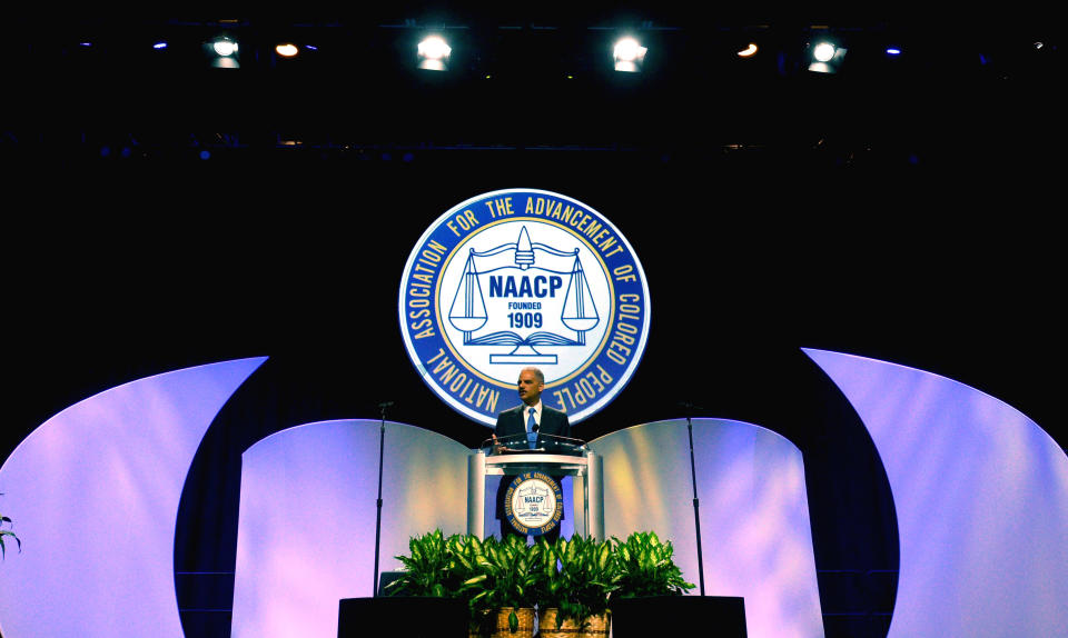 Then-Attorney General Eric Holder speaks at a podium. Behind him is a large NAACP logo.