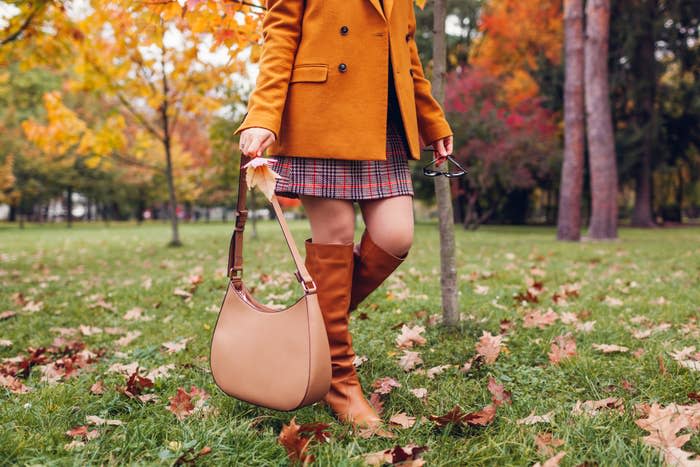 Person wearing a plaid skirt, orange coat, and knee-high boots, holding a large tan handbag and sunglasses, standing in a park with autumn leaves and trees