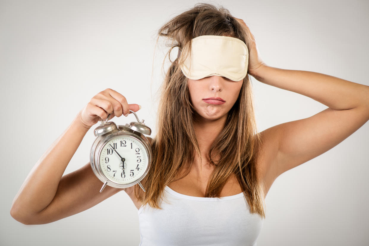 Beautiful young woman with sleeping mask holding alarm clock. She is tired and lazy in morning.