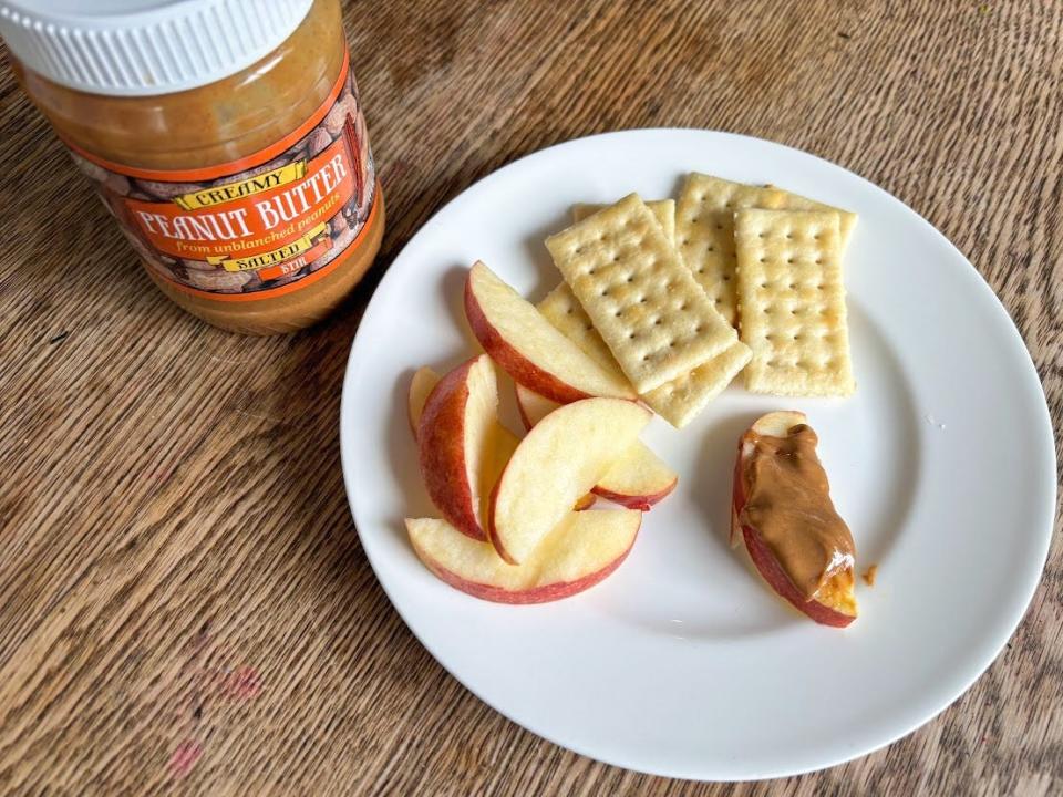 A jar of peanut butter with an orange label and white lid sits next to a plate of apple slices and crackers. Creamy-looking peanut butter is spread on one apple slice