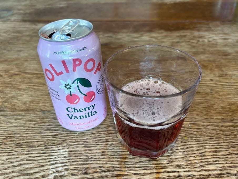 An open can of cherry-vanilla Olipop next to a small, clear glass with brown liquid inside. Both are sitting on a wooden table.