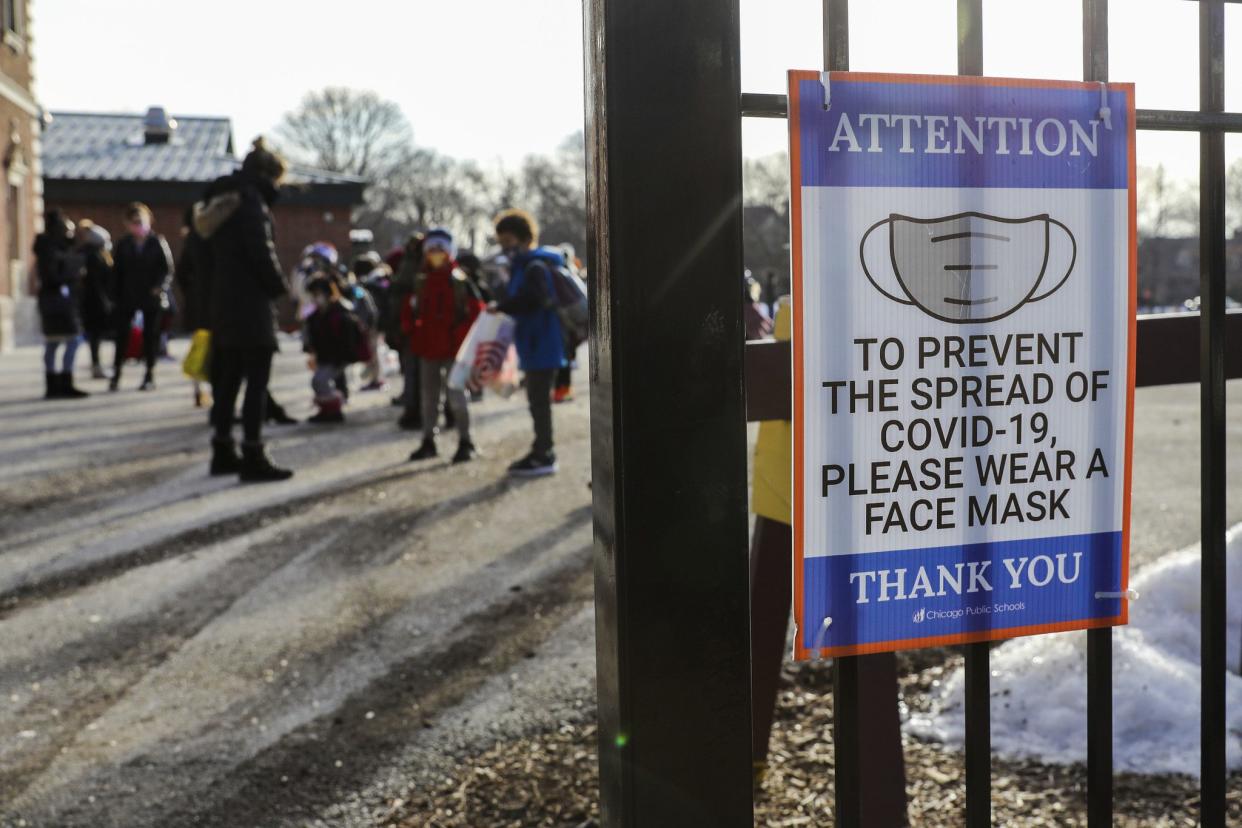 Seen through a school gate, children assemble on the playground, with a sign posted saying: Attention! To prevent the spread of COVID-19, please wear a face mask. Thank you.