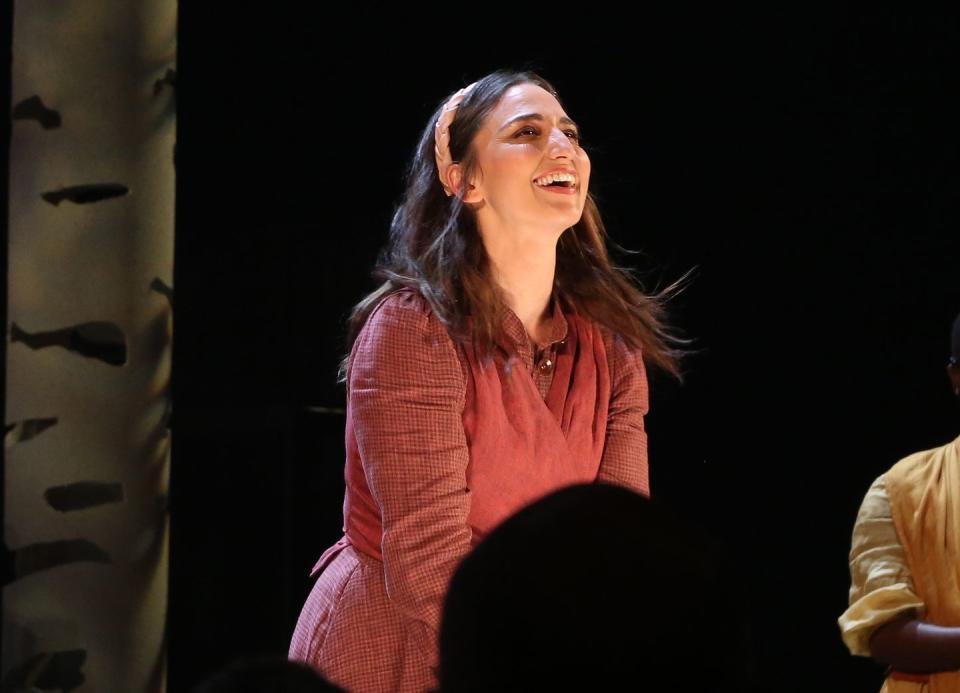 sara bareilles wearing a red outfit, smiling and looking up while standing on a stage with a dark backdrop