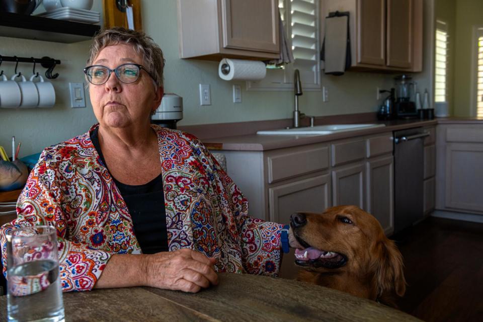 A woman with short hair, wearing glasses, is seated at a kitchen table, petting a golden retriever next to her