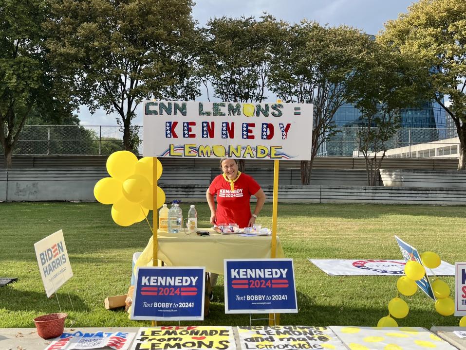 A woman at a lemonade stand hands out lemonade behind signs that read: Kennedy 2024.