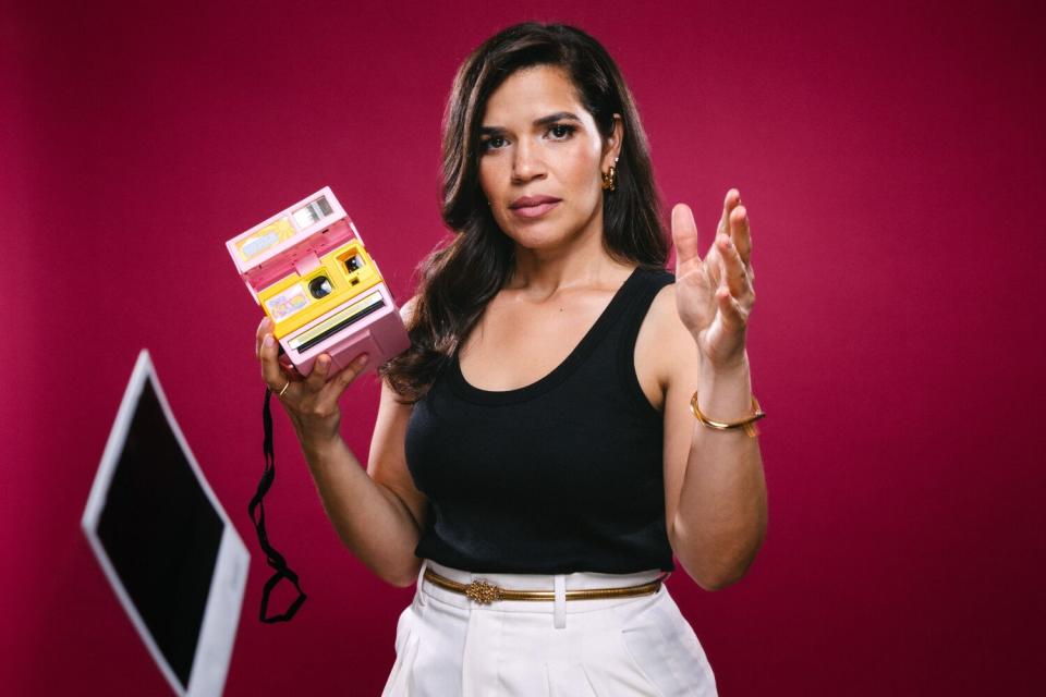 A woman in a black tank top plays with a toy Polaroid camera.