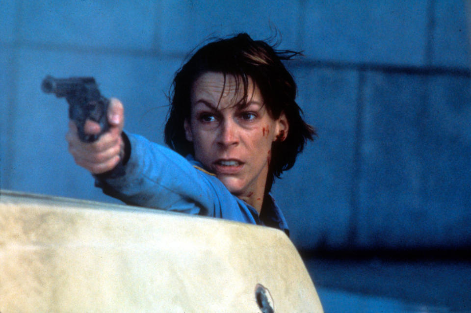 Jamie Lee Curtis hides behind a car while pointing a gun