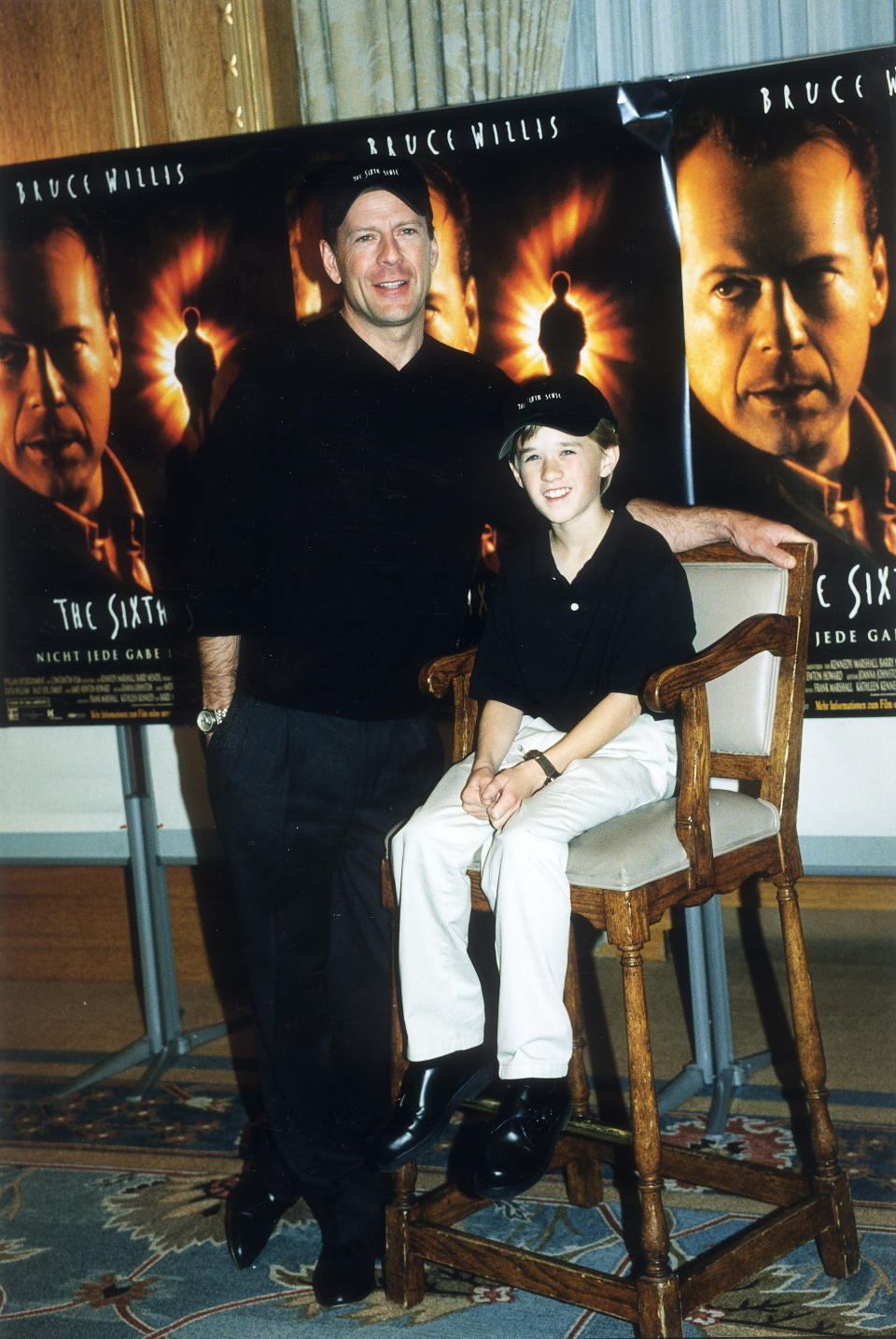 Bruce Willis stands next to a seated Haley Joel Osment at a promotional event for The Sixth Sense