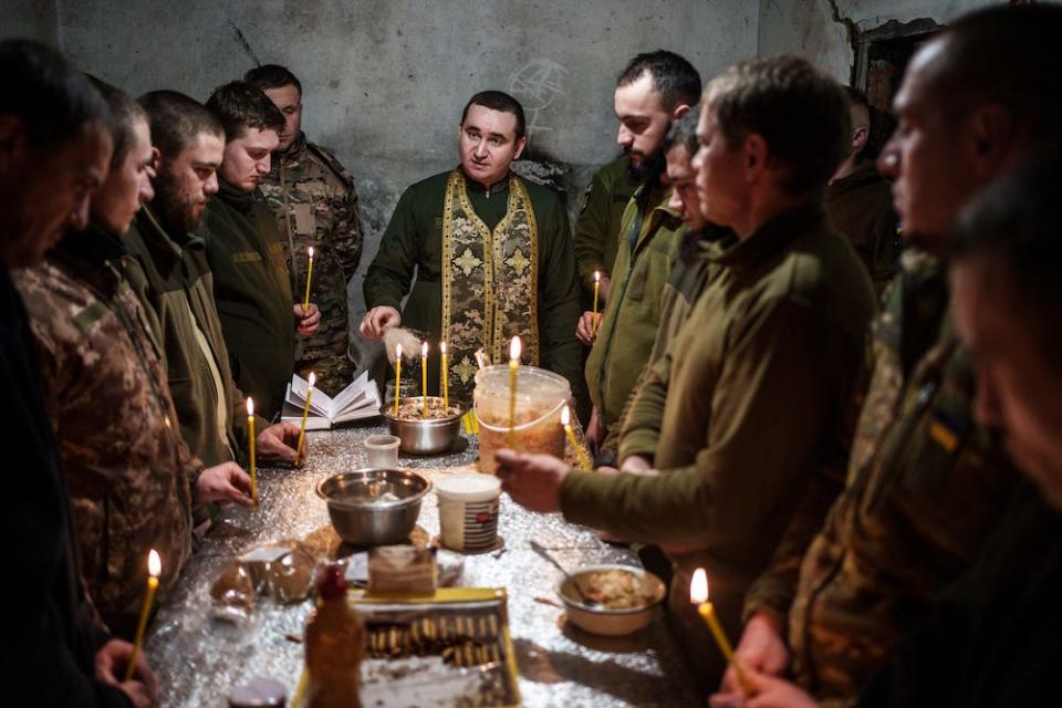 Chaplain Ivan from the 72th Brigade of the Ukrainian Armed Forces conducts a service on December 24, 2023 in Donetsk Oblast, Ukraine. He is seen standing at the head of a table surrounded by soldiers in a concrete room, some holding taper candles around basic foodstuffs.