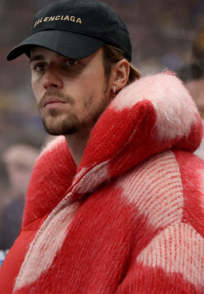 Justin Bieber in a red fluffy jacket and a black cap, looking serious