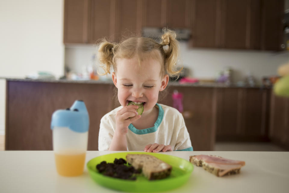 A young child with pigtails sits at a table, smiling and eating a green vegetable. A plate with food and a cup of juice are in front of her