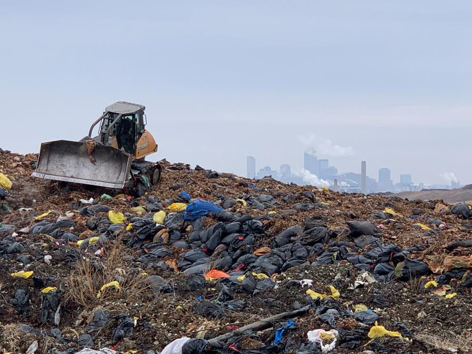 Bags of leaves that have been delivered to South Side Landfill for compost sit in a pile, waiting to be processed. A bulldozer moves the leaves around, with a view of the Indianapolis skyline in the background.
