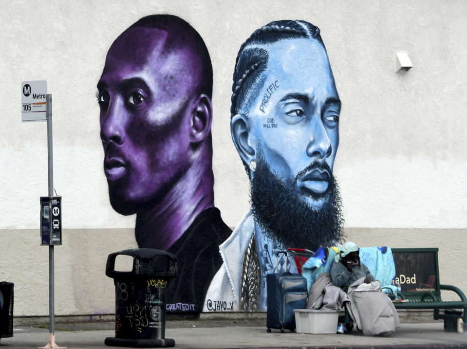 A woman sits at a bus stop on a cold day in front of a Kobe Bryant and Nipsey Hussle mural along La Cienega Blvd. in Los Angeles, Wednesday, Jan. 27, 2021. (Keith Birmingham/The Orange County Register/SCNG via AP)