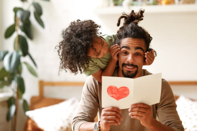 Surprise for daddy. Cute smiling afro american kid son giving father postcard with red drawn heart and covering his eyes with hands, happy child congratulating dad during family holiday at home - Credit: Getty Images