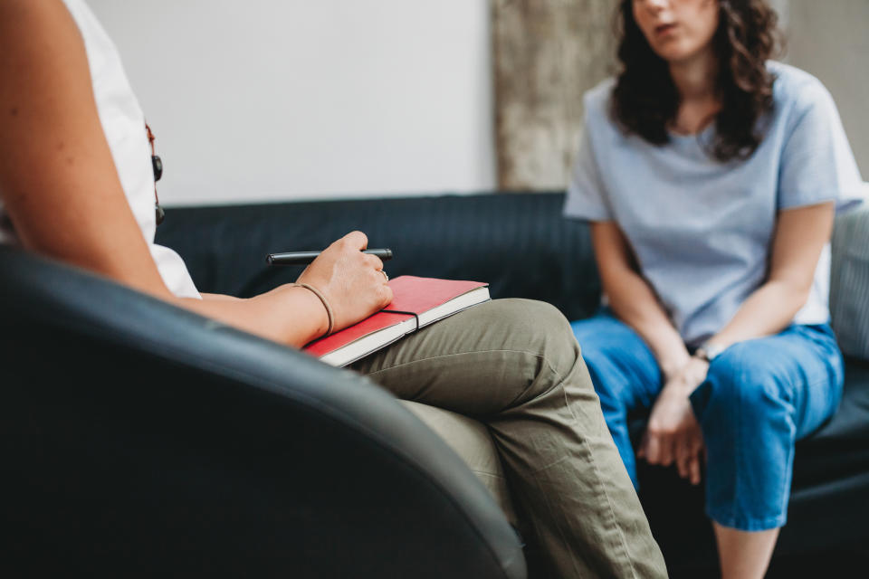 A therapist holds a red notebook and pen during a counseling session with a woman seated on a couch, discussing mental health