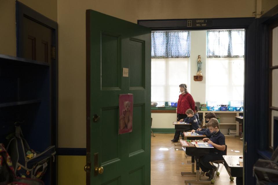 A teacher, wearing a a protective face shield, stands beside a row of students in a classroom.