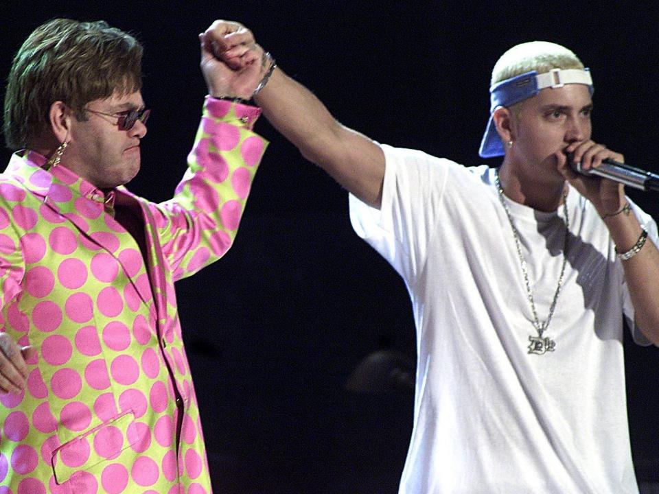 Elton John in a yellow suit jacket with large pink circles holding his hand up to hold Eminem's hand, whose wearing a white t-shirt, backwards hat, and a speaking into a microphone.
