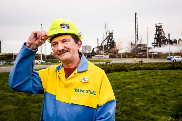 Tata Steel worker dressed in red and blue boiler suit touches his hard hat, standing in front of steelworks