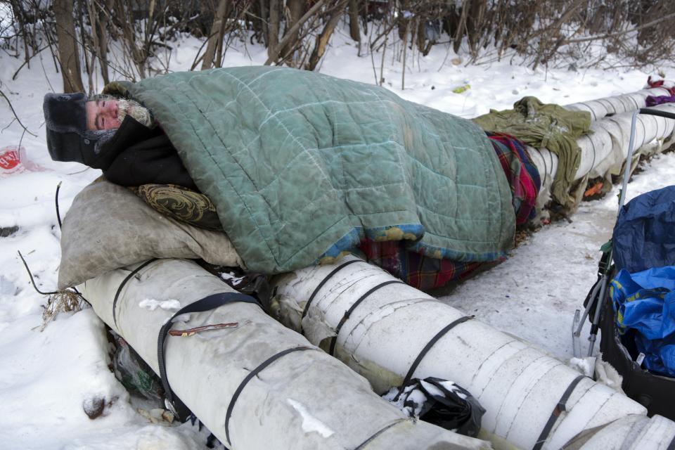 Un homme barbu portant un chapeau noir est allongé sous une couverture bleue tachée et sur de gros tuyaux