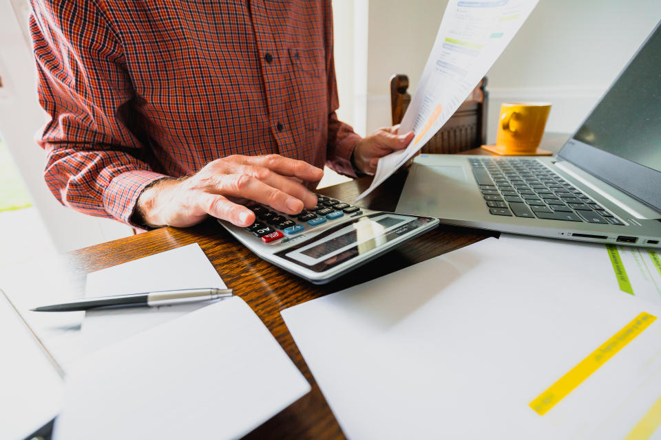 Wide angle image depicting a senior man's hand holding an energy bill while the other hand checks the numbers on a calculator. The table is strewn with documents and also a laptop. Room for copy space.