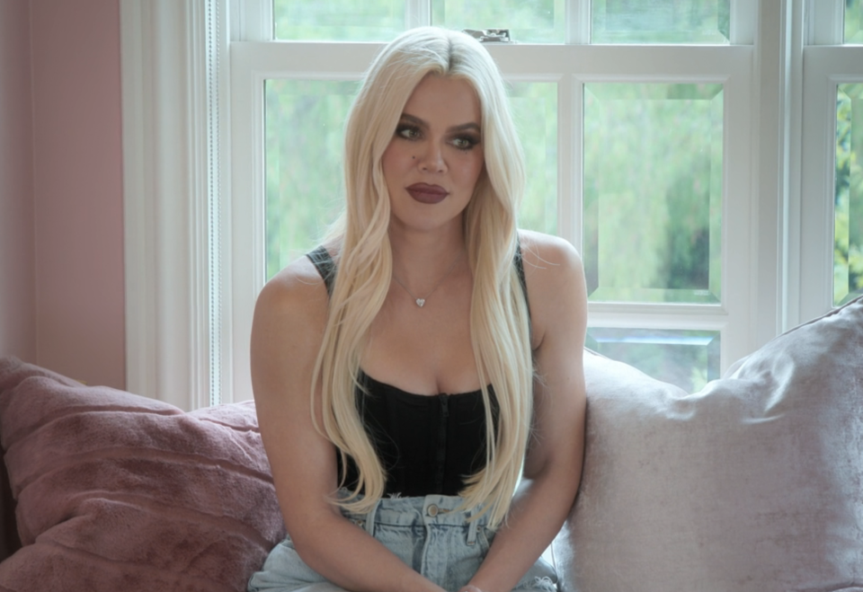 Khloé Kardashian sits on a window seat wearing a black top and jeans, with long blonde hair styled straight, looking towards the camera