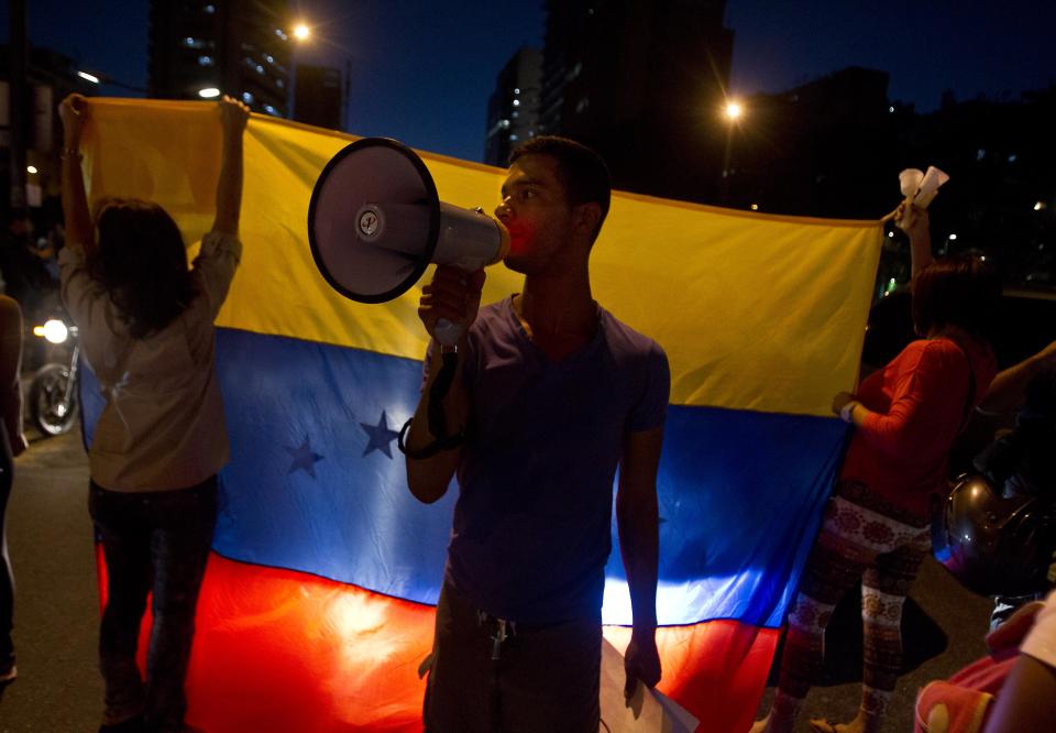 Anti-government demonstrators shout slogans against Venezuela's President Nicolas Maduro in front of Venezuela's flag in Plaza Altamira in Caracas, Venezuela, Tuesday, March 18, 2014. Protesters blocked the streets only when traffic lights turned red under the watchful gaze of the National Guard. Security forces have taken control of the plaza that has been at the heart of anti-government protests that have shaken Venezuela for a month. (AP Photo/Esteban Felix)