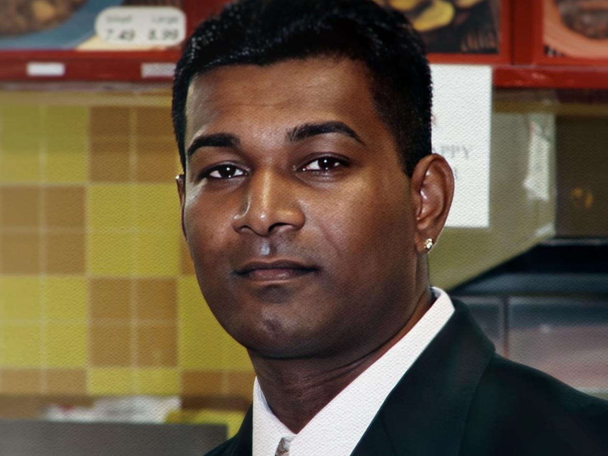 A man with dark hair and an ear piercing wearing a suit is stood in a restaurant.