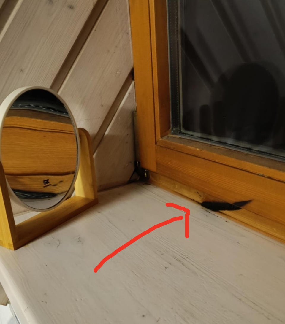 A small mirror on a wooden stand sits on a windowsill next to a closed window. There is a burn mark on the windowsill caused by the mirror