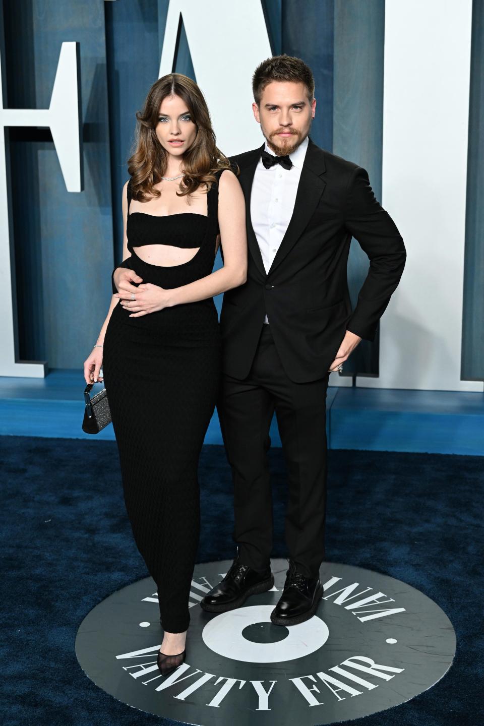 Barbara in a bandeau style dress with shoulder straps and a cut out under her chest. She's wearing a black mini purse and pointed sheer black heels. Dylan is wearing a black tuxedo, bowtie, and chunky shoes.