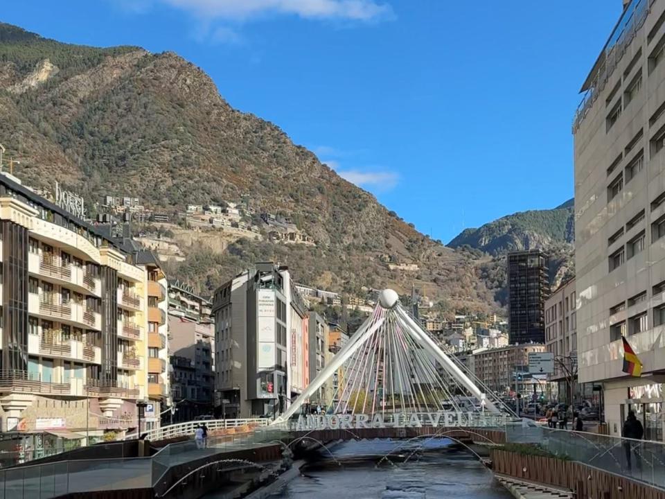 A bridge over a river with the words "Andorra La Vella" on it. There are buildings on both sides of the river and tall mountains in the background.