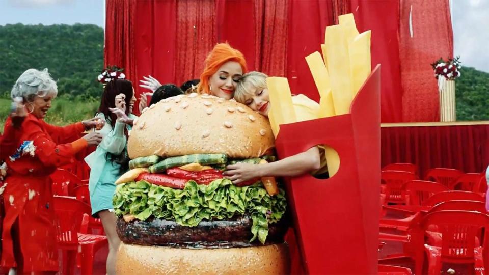 Katy Perry, dressed as a hamburger, and Taylor Swift, dressed as a box of French fries, hug each other at an outdoor event with red decorations and chairs