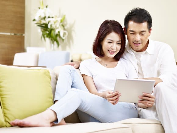 Asian couple relaxing on a couch while looking at a tablet.