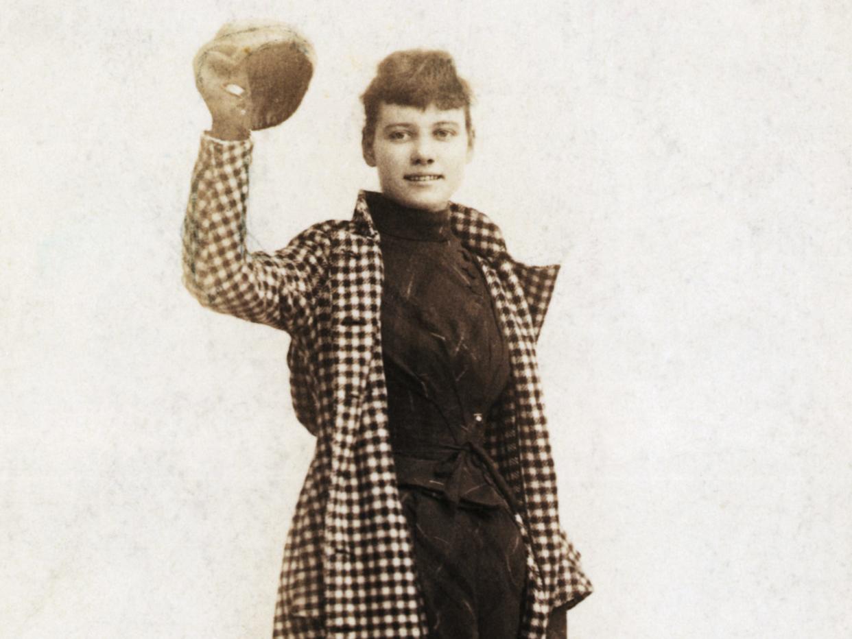 Nellie Bly holds a hat and a bag and wears a long checked jacket and dark dress