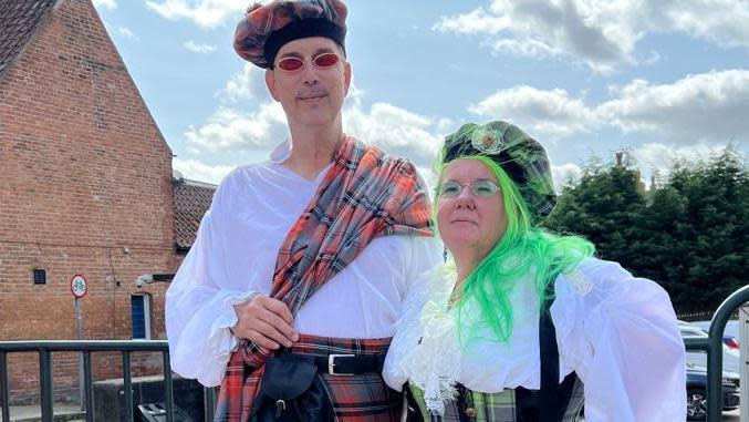 Man with red kilt and white shirt wearing a tartan beret next to woman with green kilt wearing a white lacey shirt with bright green hair