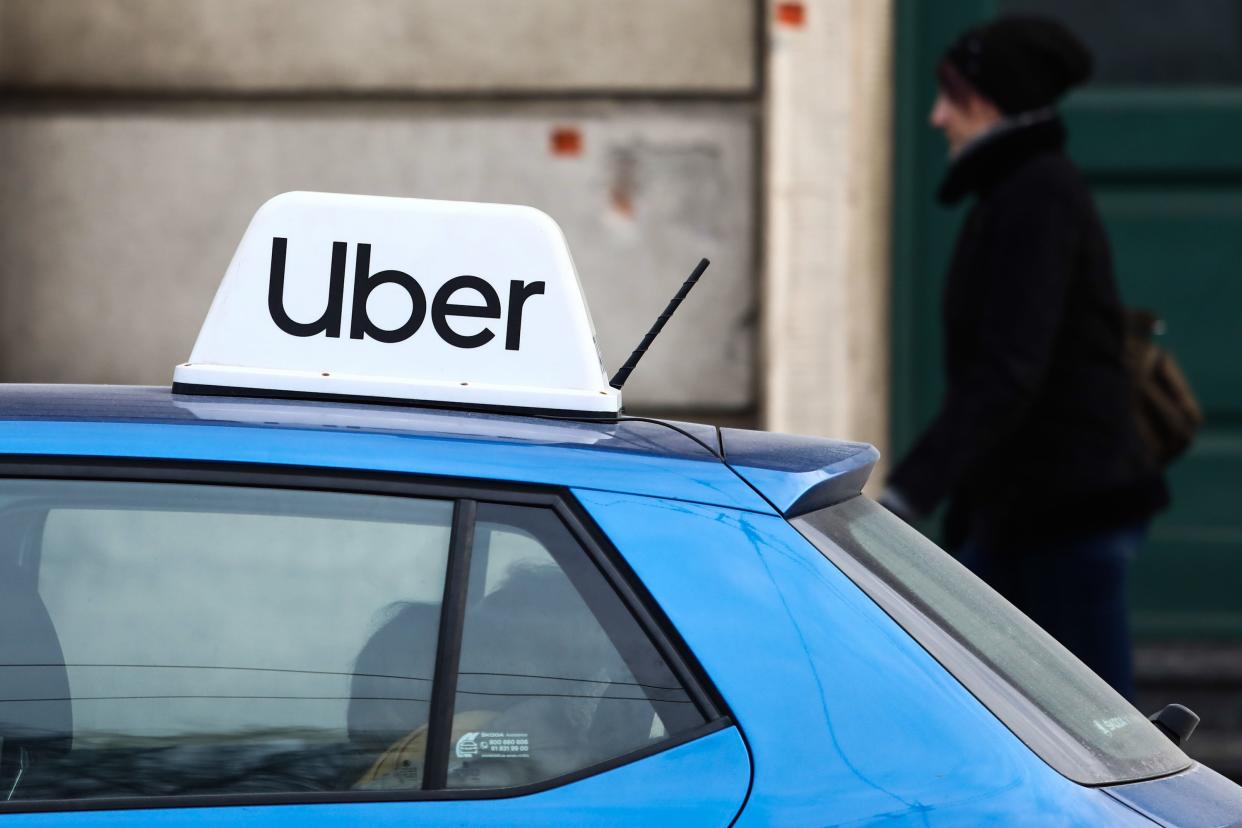 An 'Uber' placard sits on top of a car roof and a person walks past in the background.