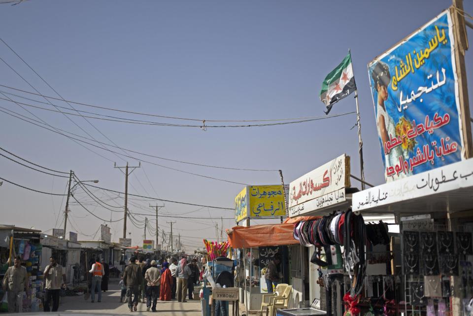 In this Thursday April 17, 2014 photo, Syrian refugees walk on the market street at Zaatari refugee camp, near the Syrian border in Jordan. A Syrian revolution flag flies above one shop, between banners with Arabic writing that read "Yasmeen al-Sham for Beauty," "Al-Mahabba accessories shop," and "Al-Refaey Jewelry shop." (AP Photo/Khalil Hamra)