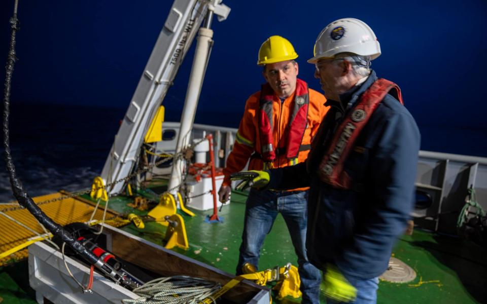 Two men in hard hats on the green-painted deck of a ship at night