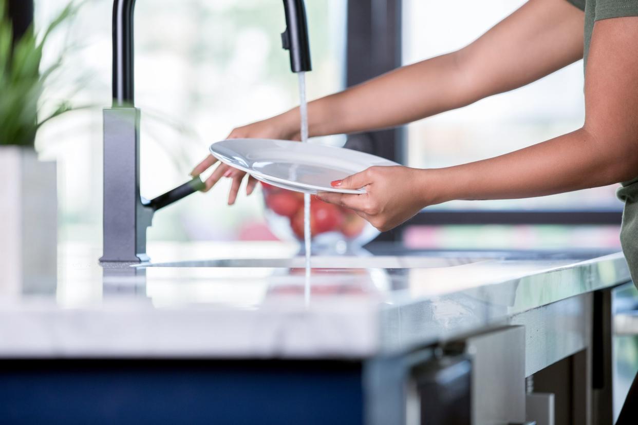 Young adult unrecognizable Hispanic woman turns on running water from faucet while washing dishes at kitchen sink.