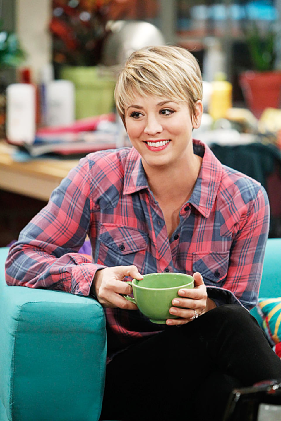 Kaley Cuoco in casual plaid shirt holding a mug, sitting on a couch in a cozy, cluttered living room setting, her hair is a short pixie cut with sideswept bangs