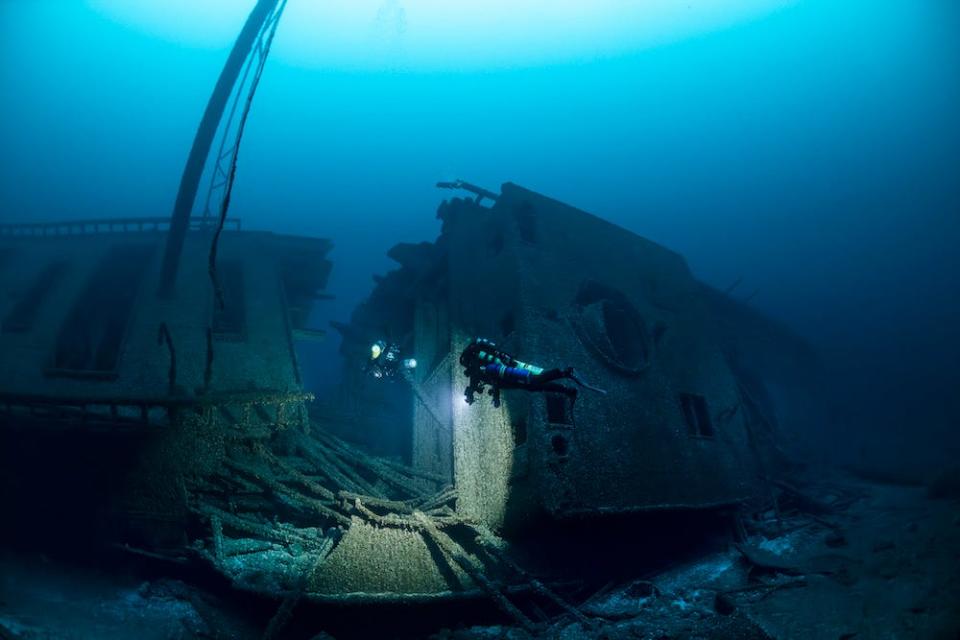 A diver floats past a massive break in the S.S. Norman in the Great Lakes, illuminating it with a torch. The image is a winner in the 2024 Underwater Photographer of the Year Awards.