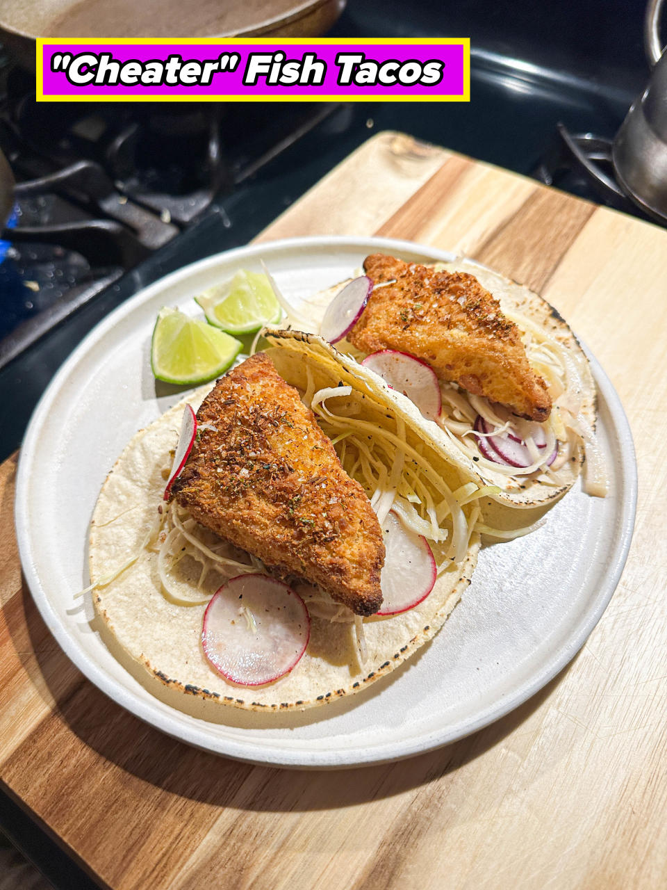 Two fish tacos served on corn tortillas, topped with shredded cheese, radish slices, and a side of lime wedges on a white plate