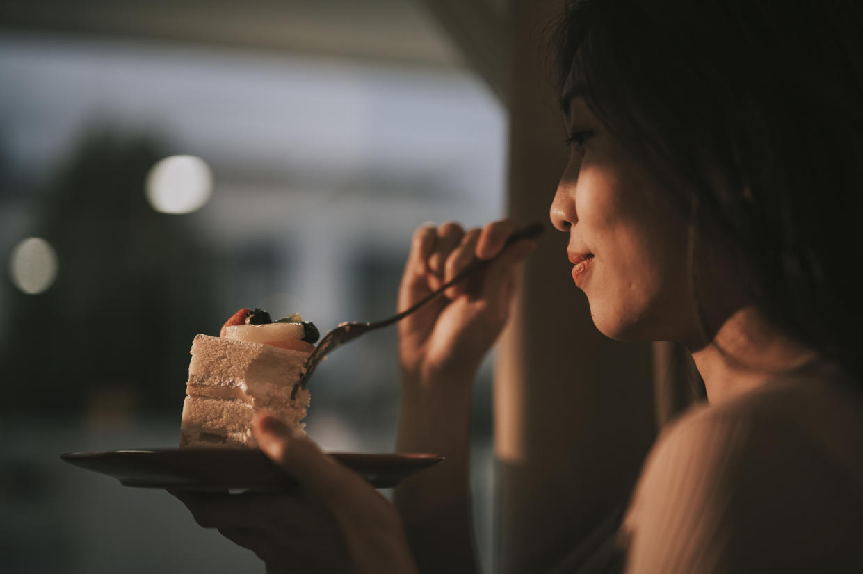 Asian Chinese beautiful woman enjoying slice of cake at home during sunset looking away through window curtain