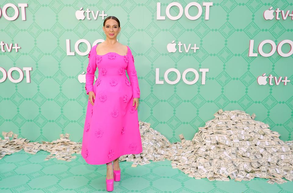Maya Rudolph in a pink dress and pink shoes in front of a pile of money