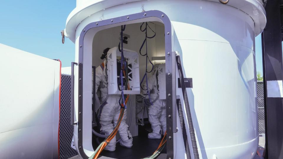 astronauts in white spacesuits stand inside a white airlock