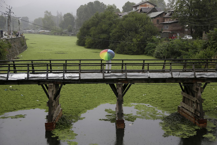 <p>Während eines Regenschauers steht ein Mann mit einem bunten Regenschirm auf einer Brücke über dem Dal Lake in Srinagar im indisch kontrollierten Kashmir und fischt. (Foto: Mukhtar Khan/ AP)</p>