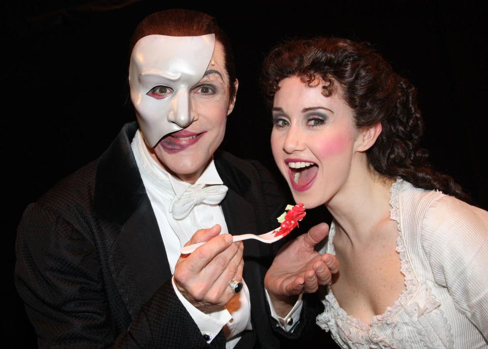 howard mcgillin and elizabeth loyacano dressed as the phantom of the opera characters, with mcgillin holding a plastic spoon with cake up to loyacano's open mouth