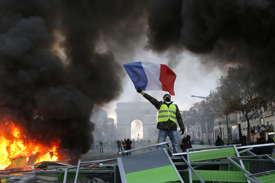 A demonstrator waves the French flag onto a burning barricade on the Champs-Elysees avenue with the Arc de Triomphe in background. Source: AP