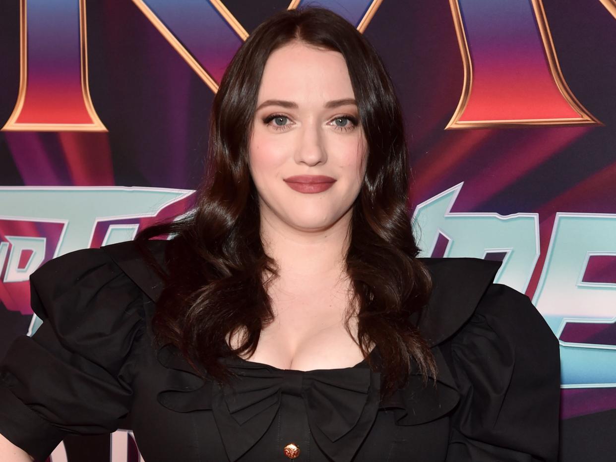 Kat Dennings poses with a hand on her hip on a red carpet.